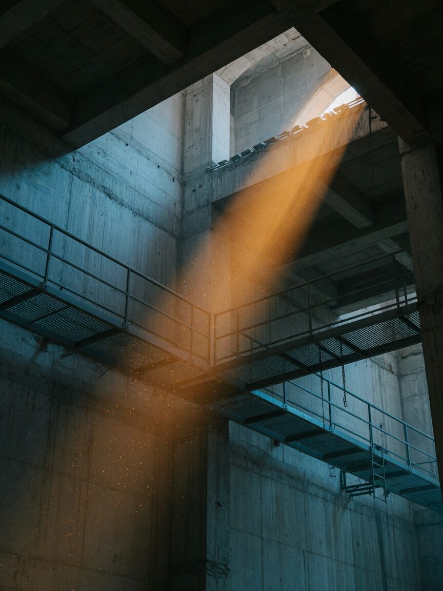 Amber shaft of light cutting through a high concrete interior with steel mesh catwalks.